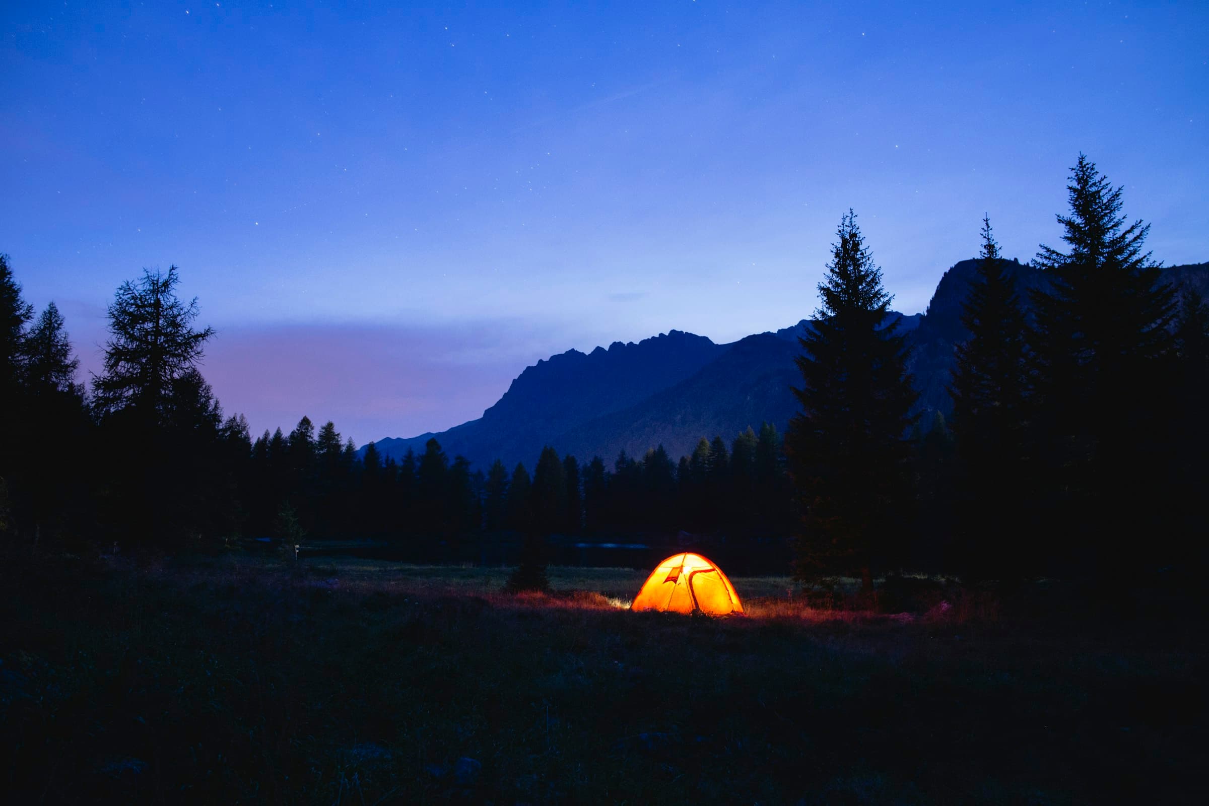Glowing tent in a meadow beneath mountains at blue hour