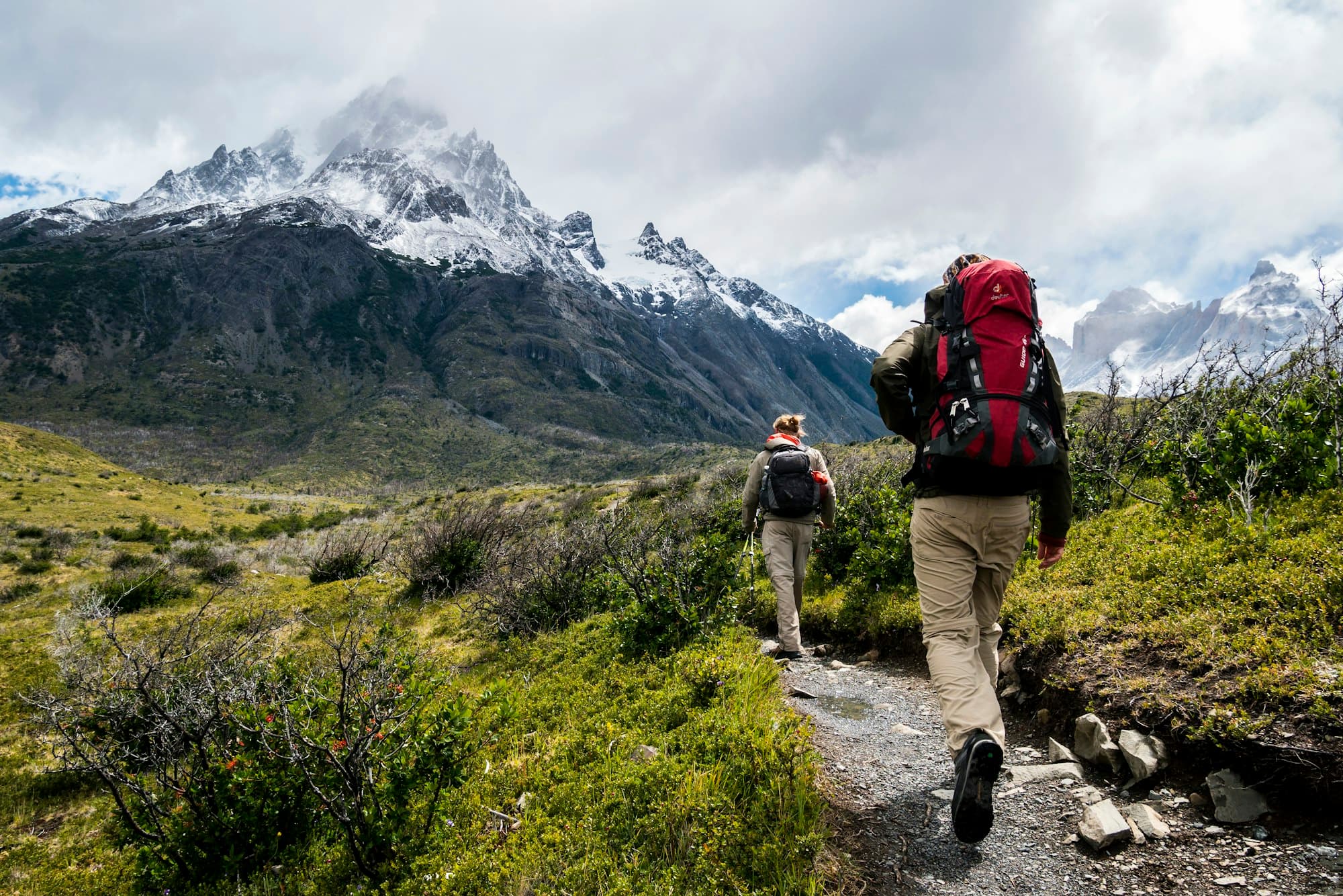 Two hikers with backpacks walking a trail toward snow-capped mountains
