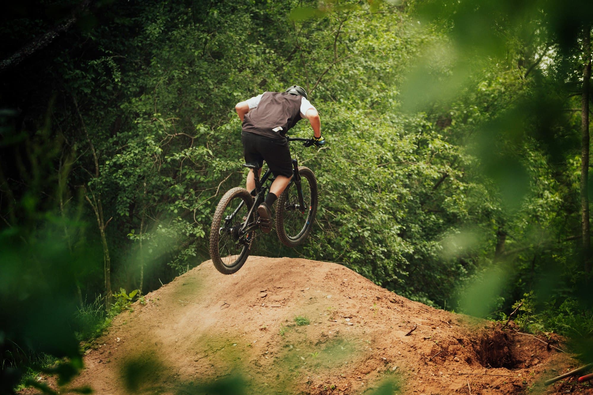 Mountain biker launching off a dirt jump in a green forest