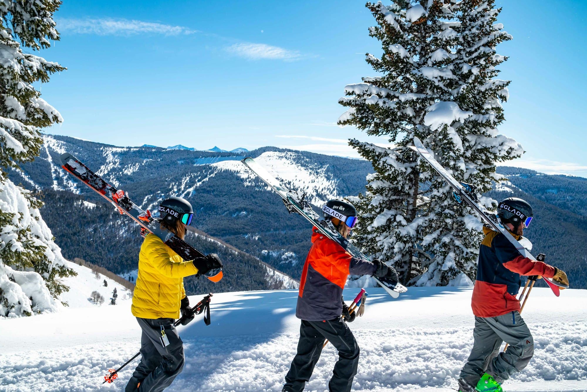 Three skiers hiking up a snowy ridge with skis on their shoulders